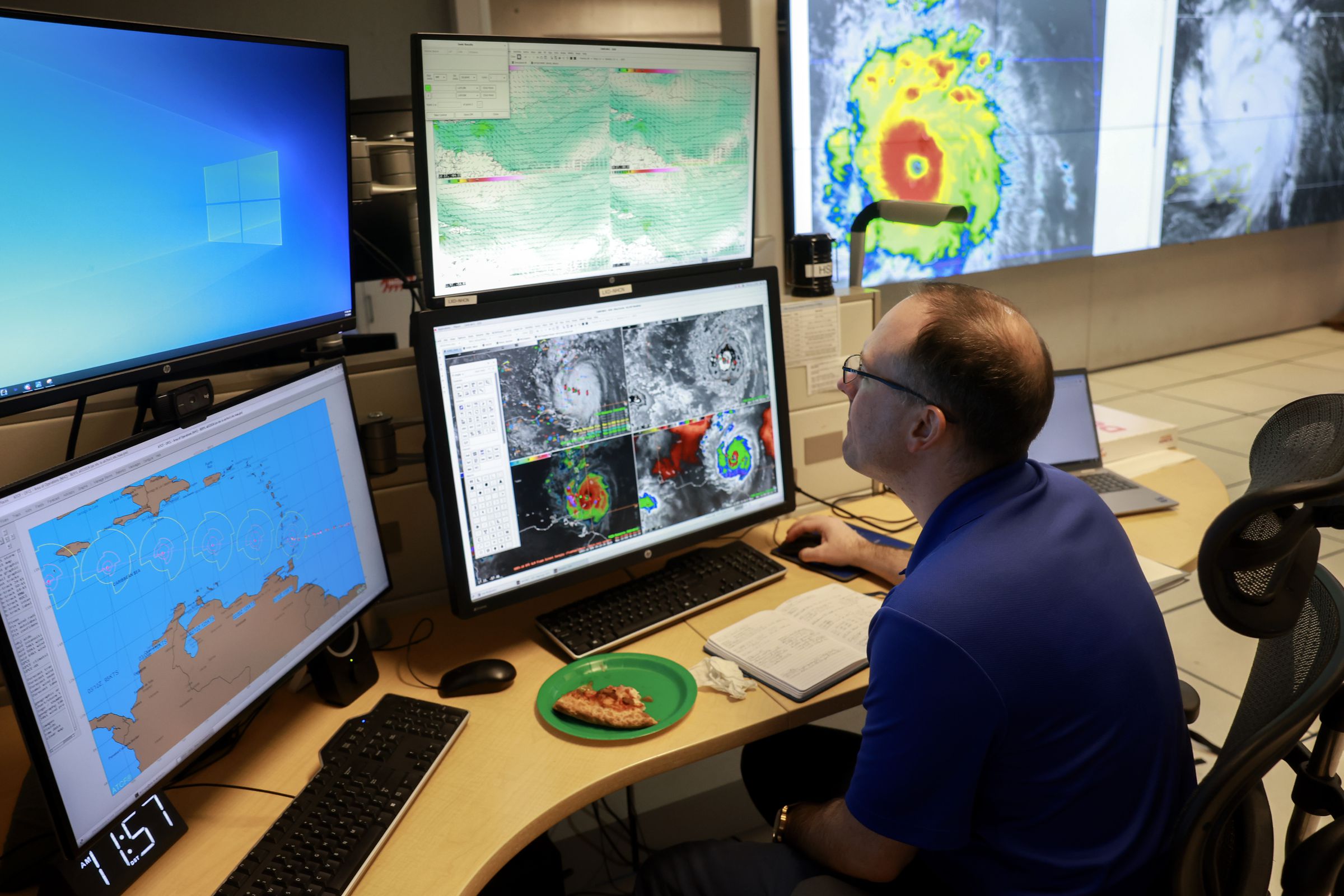 A person sits at a desk, surrounded by computer screens and wall projections of hurricane activity.