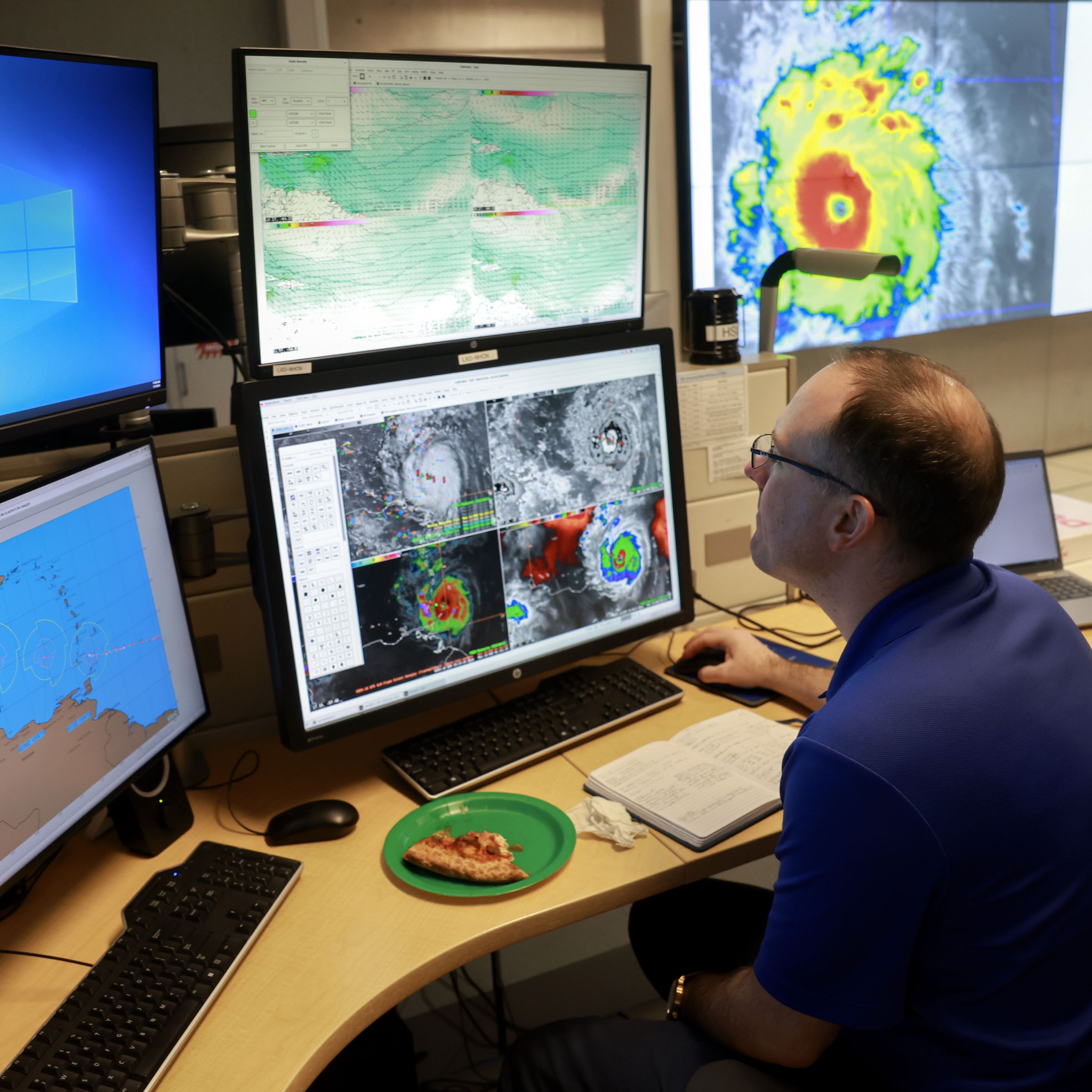 A person sits at a desk, surrounded by computer screens and wall projections of hurricane activity.