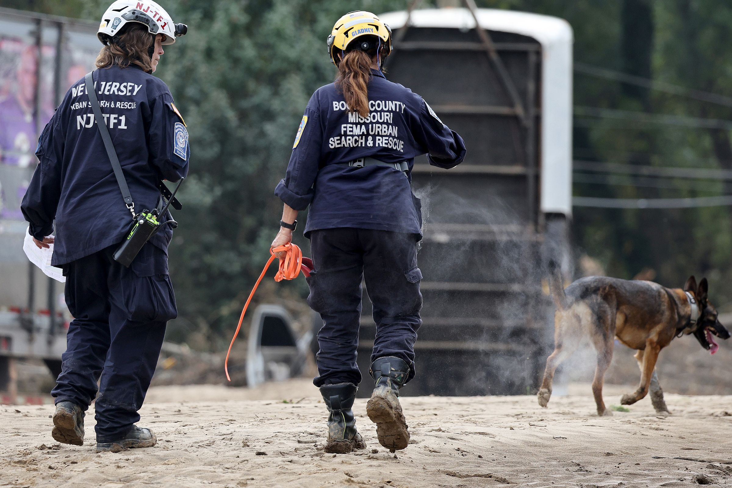 Two people wearing FEMA uniforms and helmets seen from behind, walking next to a dog.