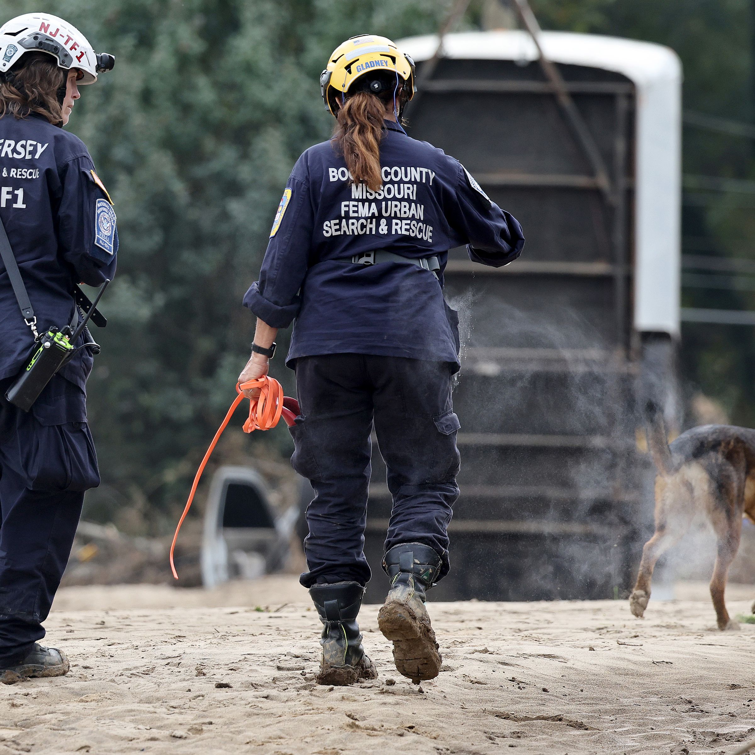 Two people wearing FEMA uniforms and helmets seen from behind, walking next to a dog.