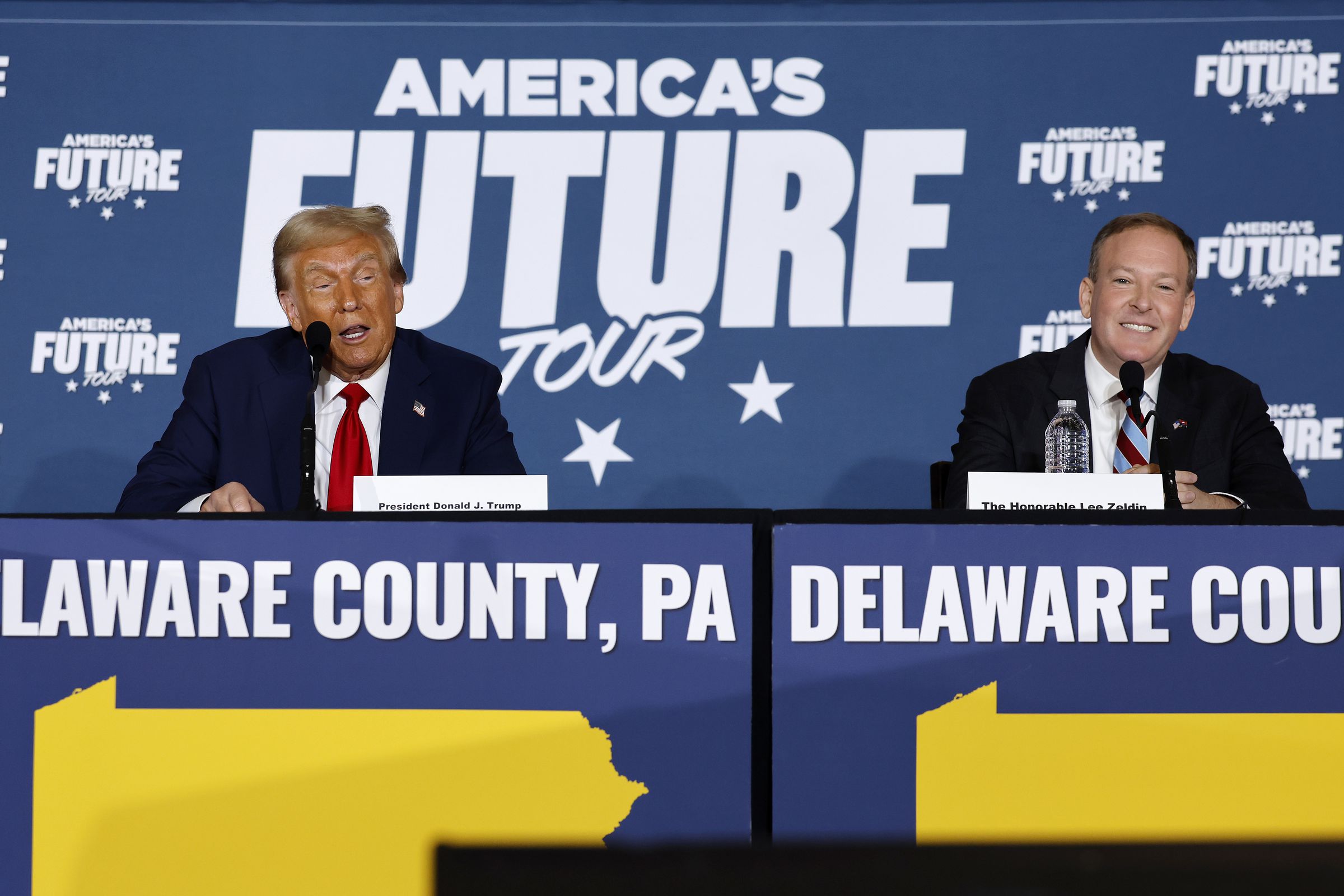 Donald Trump and Lee Zeldin sit next to each other on a panel. A sign behind them says “America’s Future Tour.”