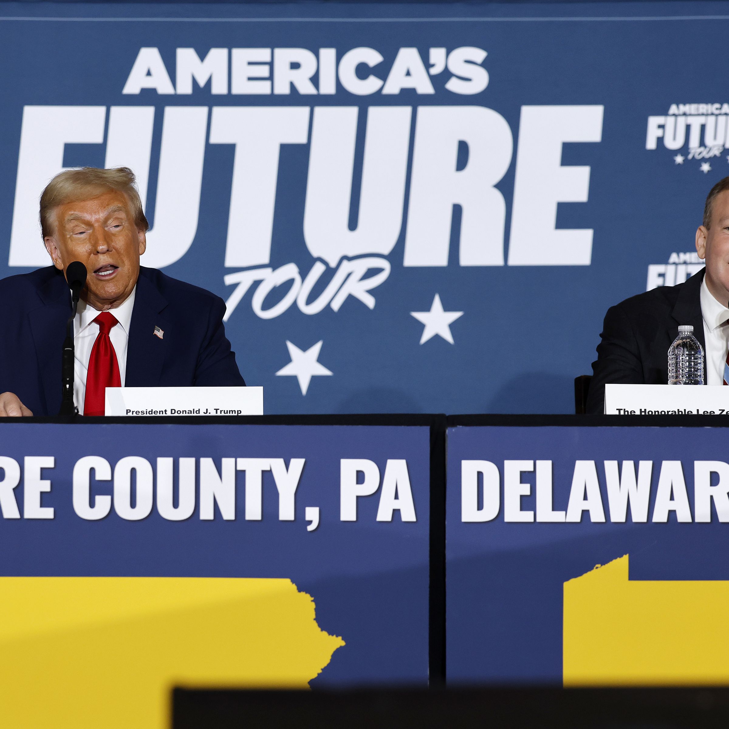 Donald Trump and Lee Zeldin sit next to each other on a panel. A sign behind them says “America’s Future Tour.”