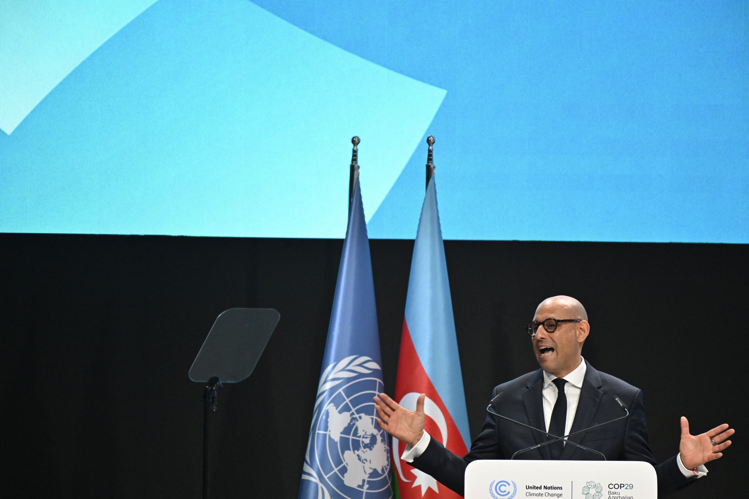 A man wearing a suit and glasses speaks at a podium in front of flags of the United Nations and Azerbaijan.