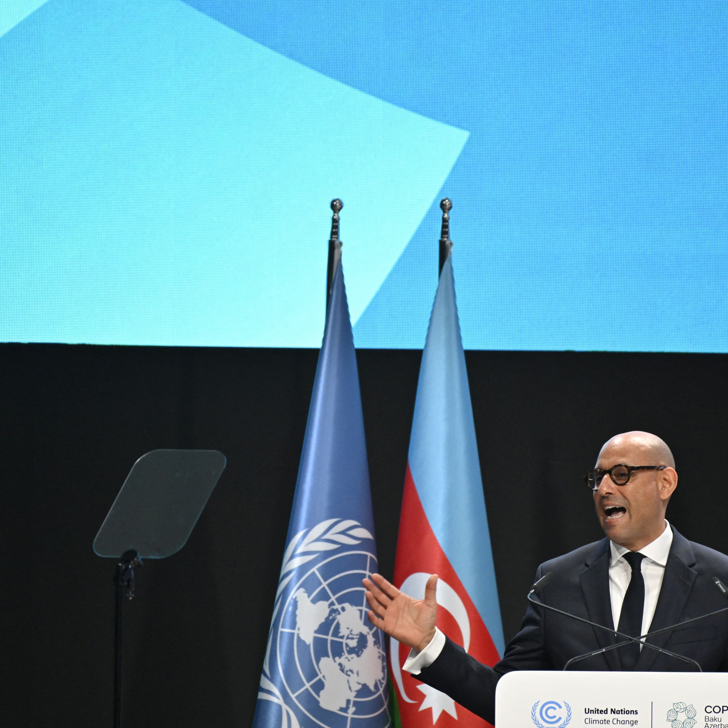 A man wearing a suit and glasses speaks at a podium in front of flags of the United Nations and Azerbaijan.