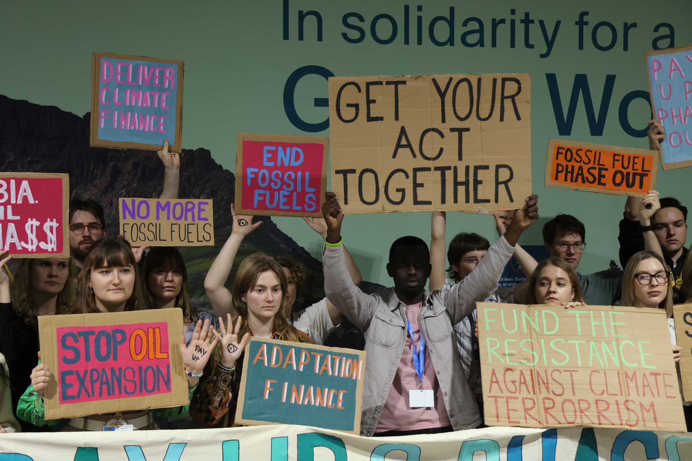 A group of protesters holding signs stand together. One person in the center holds up a sign that says “get your act together.”