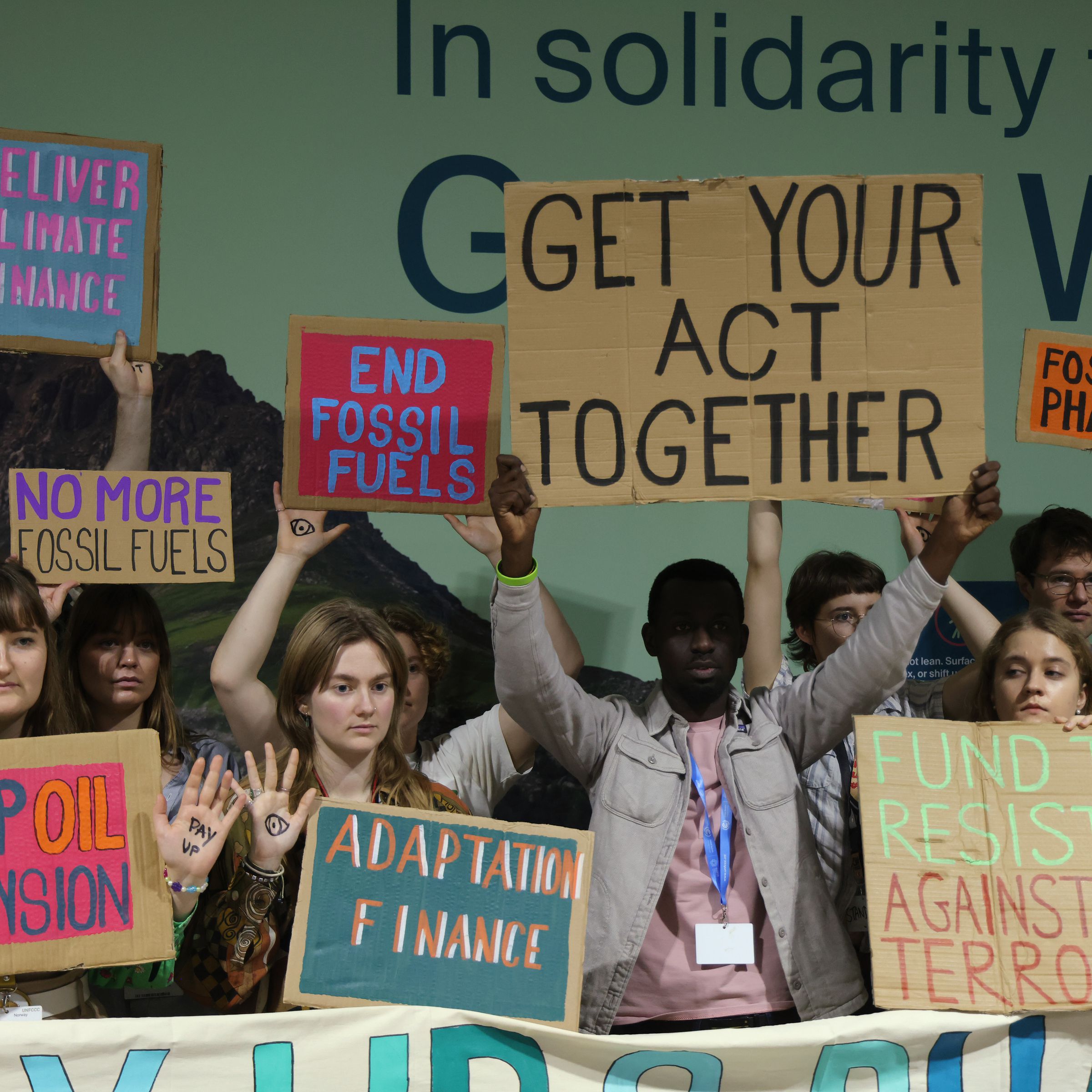 A group of protesters holding signs stand together. One person in the center holds up a sign that says “get your act together.”