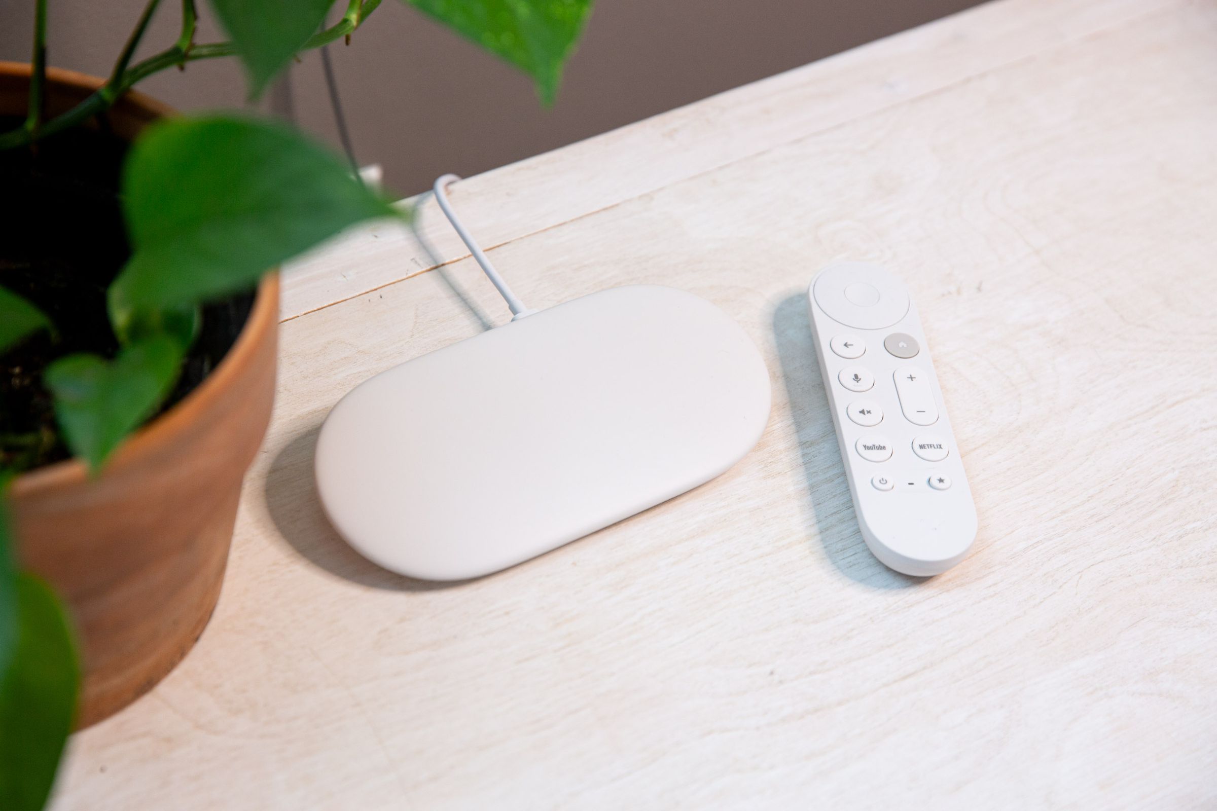 A white Google Streamer device next to a white remote on a white TV console next to a green plant.