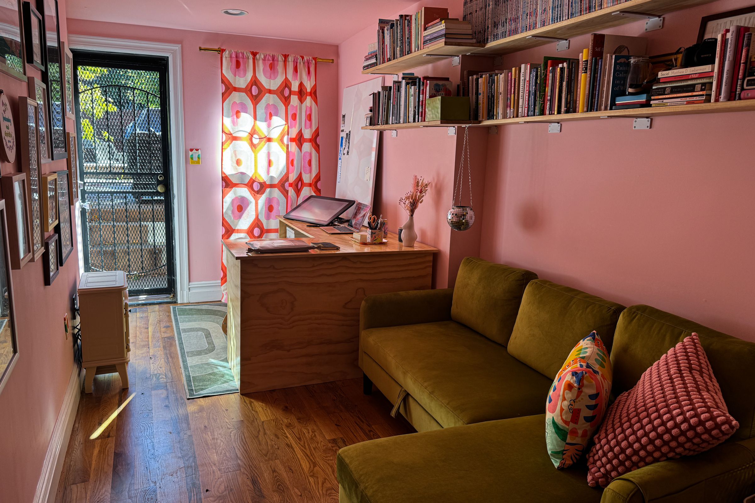 Pink home office with door to outside at far end, desk and couch on right side, photos on wall at left.