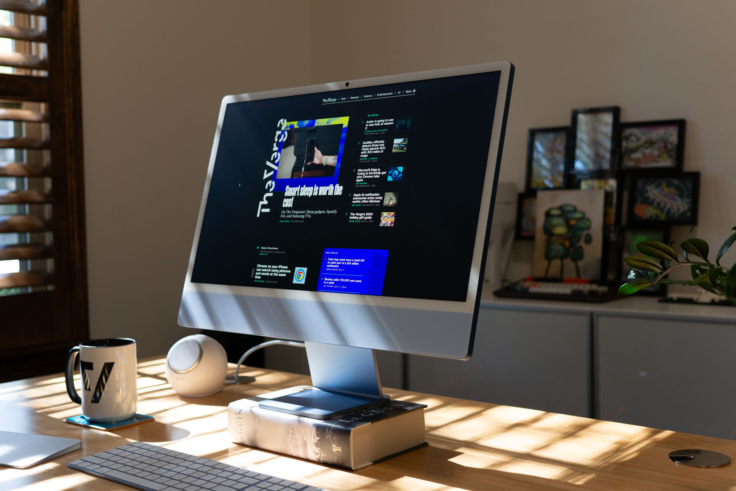 A blue all-in-one computer on a sun-lit bamboo desk. It is sitting on a hardcover book called “the unreal and the real” by Ursula K LeGuin.