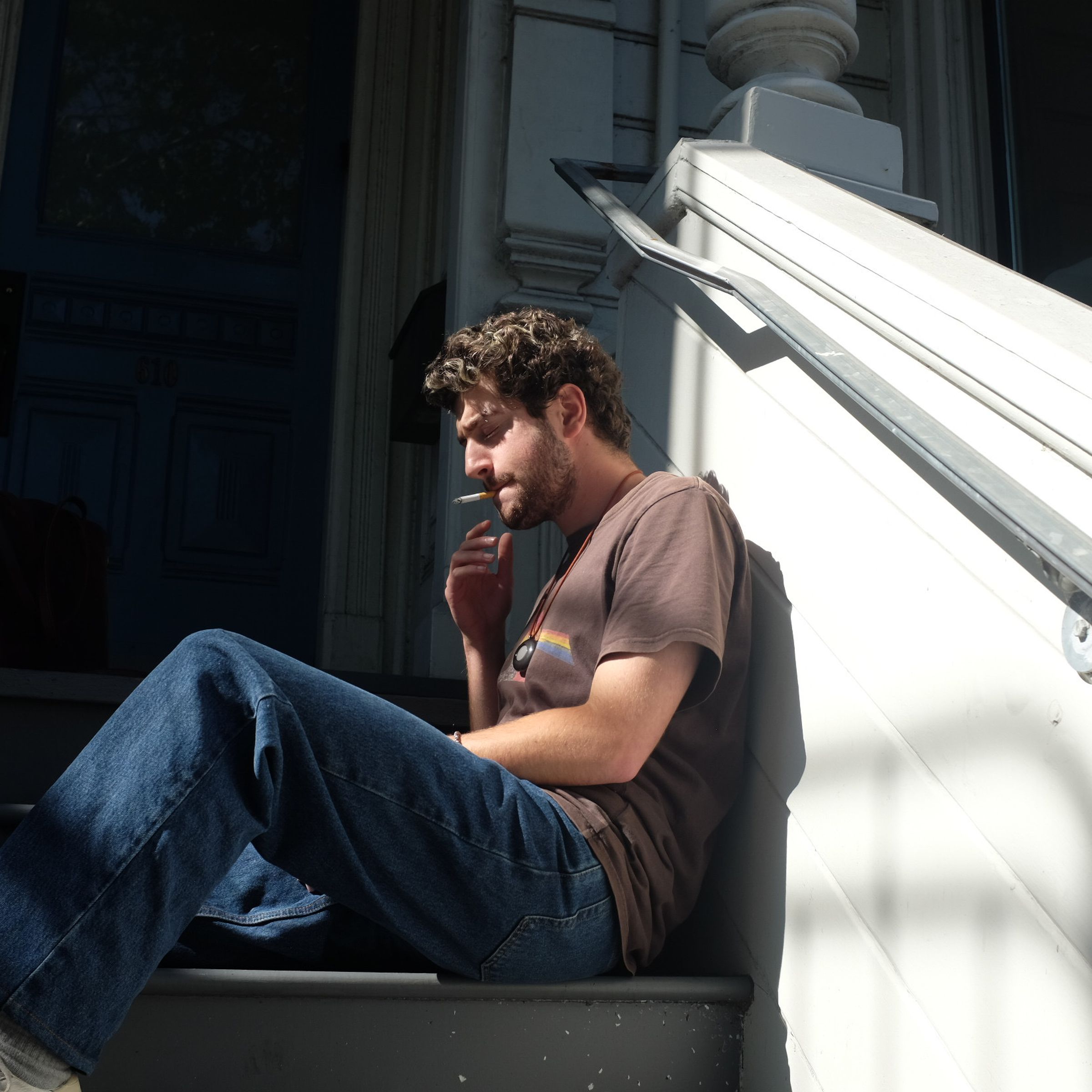 Avi Schiffmann sitting on the steps of a building, smoking a cigarette, as his Friend device lies on his chest.