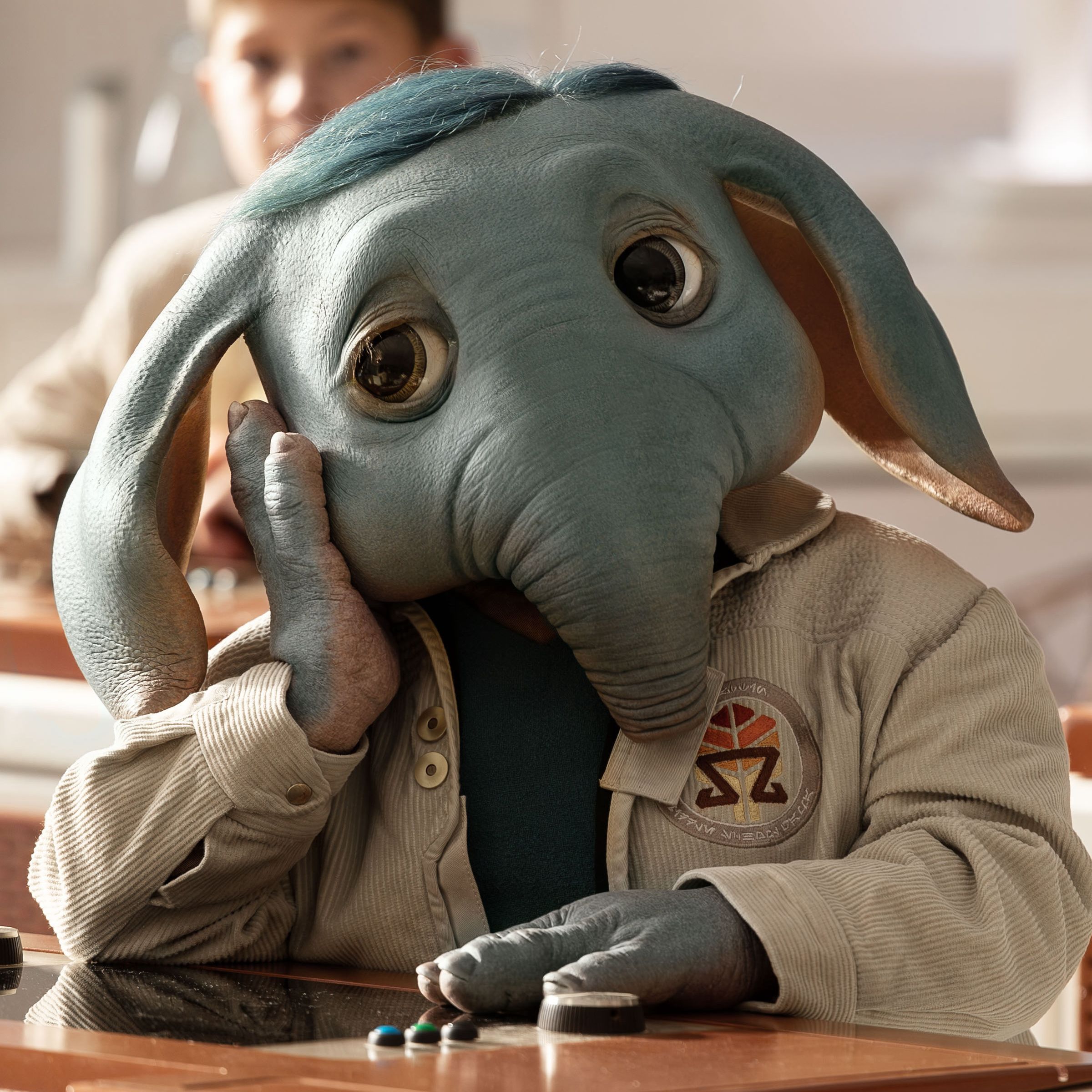 An anthropomorphic elephantine alien wearing a cream-colored jacket and sitting at a desk. The alien is resting its head on its cheek and looking to its right with an exasperated expression.