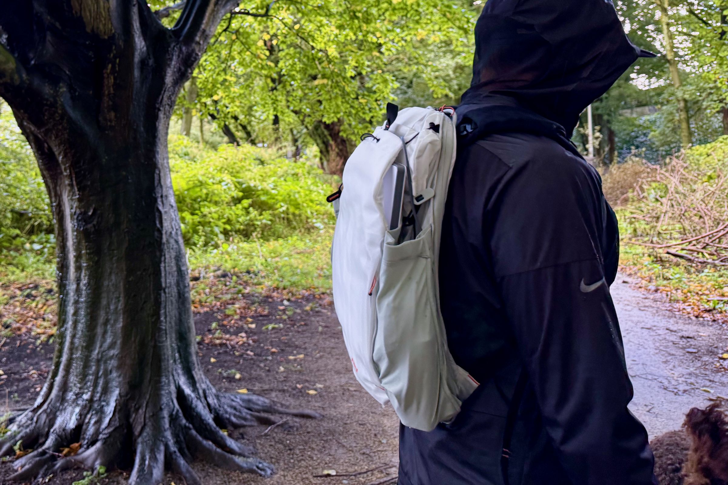 The Outdoor Backpack worn outside in profile next to a damp tree with lots of green foliage in the background.