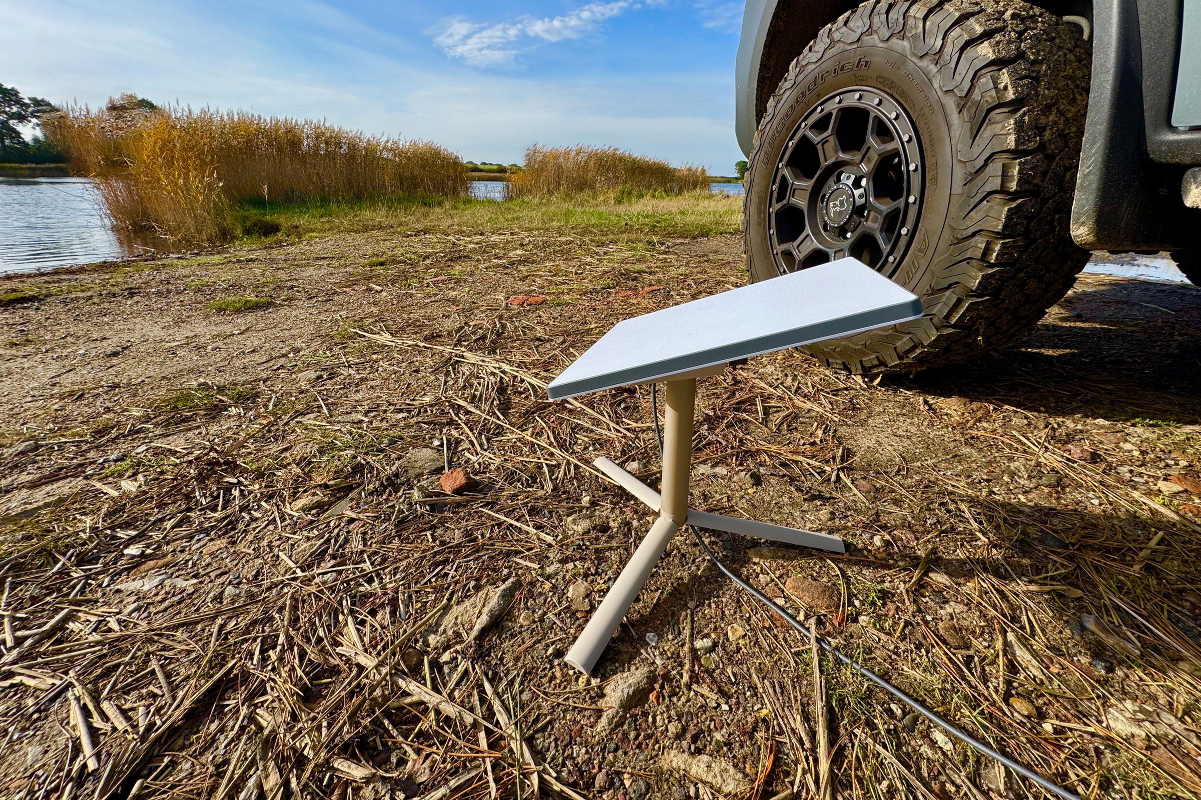 The Starlink Mini dish mounted on a small tripod using the included pole mount. It sits on the dirt in front of a pond with a large 4 x 4 van tire, reeds, and blue sky visible in the background.