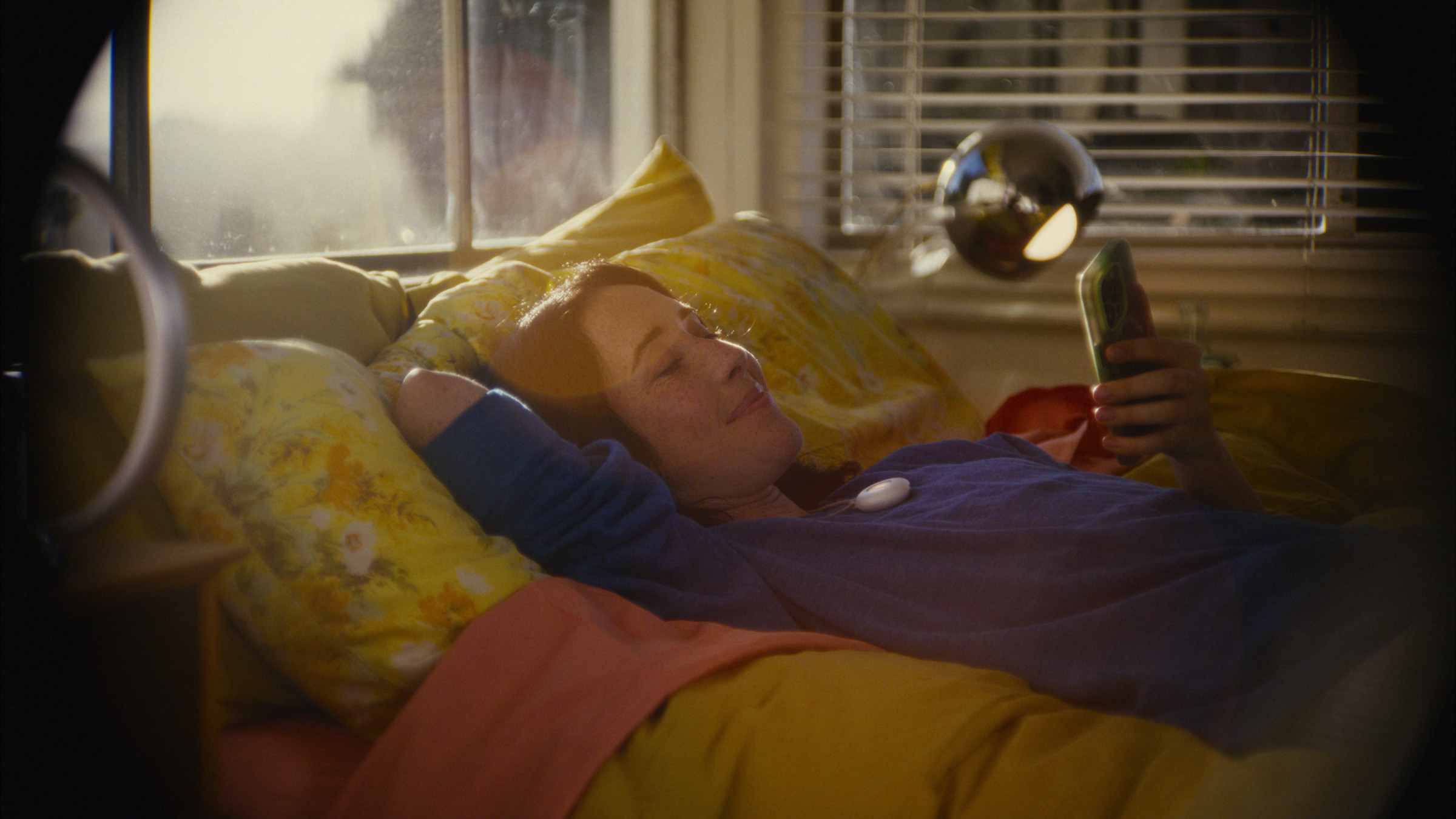 A woman lies in a bed with yellow blankets, she wears a long sleeve blues shirt rolled up to her elbows. On her chest is the Friend pendant, and she holds her phone to view its messages.